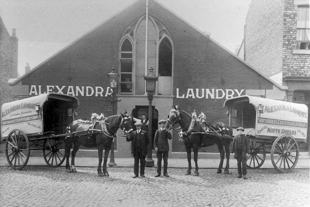 Photo of the baths building taken around 1925, many years after the baths closed. It is now occupied by Alexandra Laundry. But the front of the long apex roofed building still has the triple pointed arched windows over the entrance.  In front of the building are two horse-drawn laundry wagons with their drivers.
