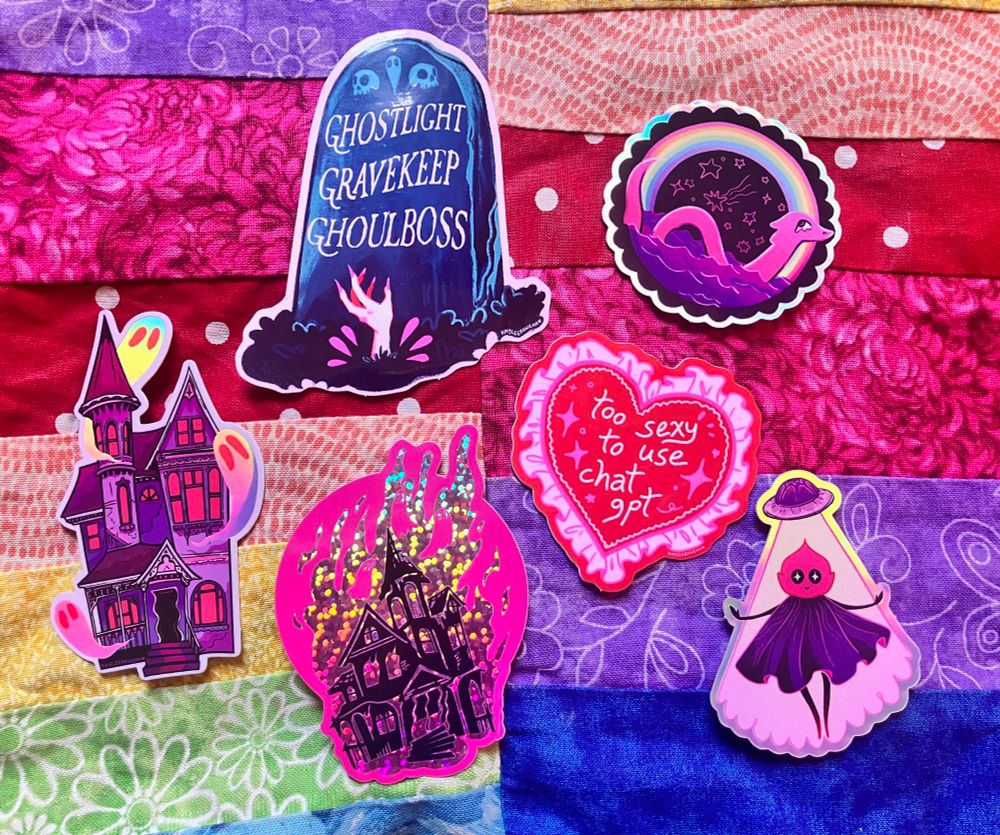 a group of stickers on a rainbow fabric background: a purple house with holographic ghosts, another house with glittery fire coming off of it and several pink ghosts in the flames, a dark teal gravestone with the words “ghostlight gravekeep ghoulboss” on it and a pink-nailed hand bursting from the ground in front of it, a frilly heart with the words “too sexy to use chatgpt” on it, and holographic stickers of the loch ness monster and flatwoods monster.