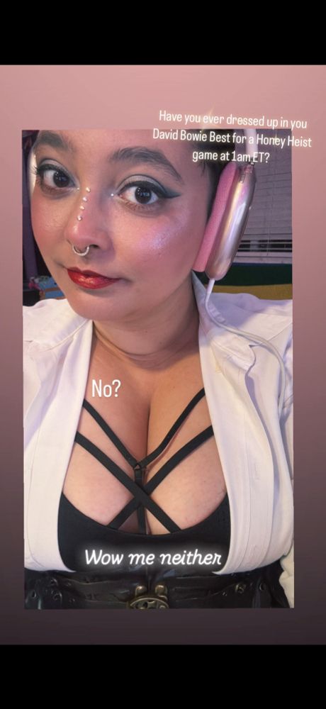 Falcon with dramatic white and black eye make up, wearing a white button up that is open to reveal a strappy bra. On it are the words "Have you ever dressed up in your David Bowie Best for a Honey Heist game at 1am ET? No? Wow me neither"