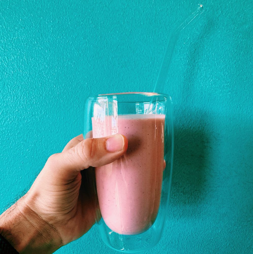 Hand holding a creamy pink beverage in a clear double-walled glass cup with a glass straw, held against a blue wall to contrast the warm tones of the drink.