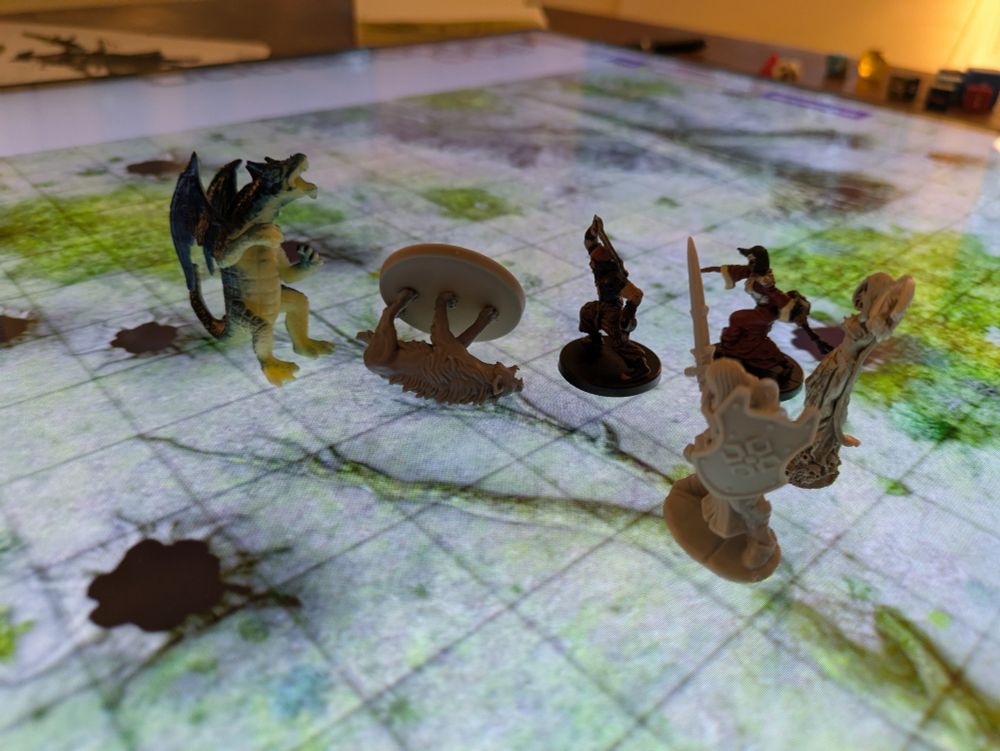 TTRPG miniatures on a lit screen map of a forest floor. A dragon miniature stands over a wolf miniature that is knocked over while the rest of the group looks on. 