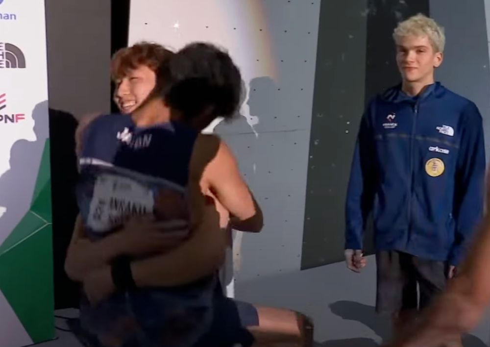 Lee Dohyun, a boulderer, picks up Anraku Sorato (another boulderer) while the Mejdi Schalk (another boulderer) watches awkwardly in the back 