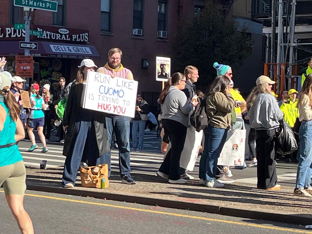A bunch of pedestrians cheering on NYC marathon runners in Brooklyn. One pedestrian is holding a "Run Like Cuomo is Trying to Hug You" sign. 