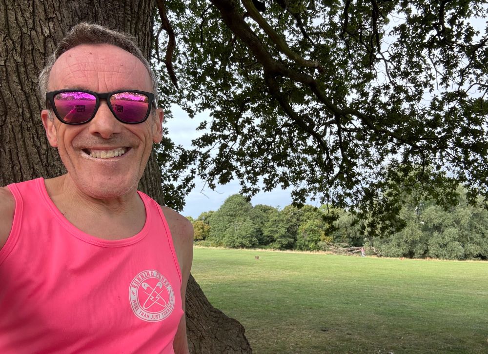 Tired looking runner with pink vest and shades, his hair is a mess - he should have taken the photo before running.  He is standing under a tree.