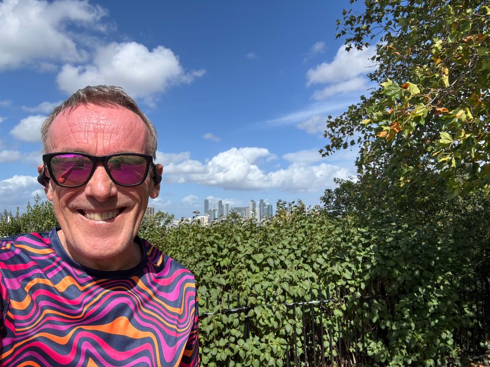 Aging man with psychedelic t shirt grins into the camera, his hair is less of a state than normal. Behind him are overgrown shrubs and behind them blue sky and the towers of Canary Wharf.