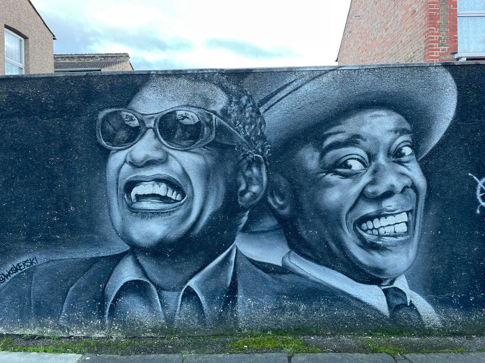 A black and white mural, made to look like a black and white photo on a fence. Ray Charles is on the left in sunglasses in an open neck shirt and jacket. Louis Armstrong appears just behind and to the right wearing suit,shirt and tie and large hat. A house is just visible above.