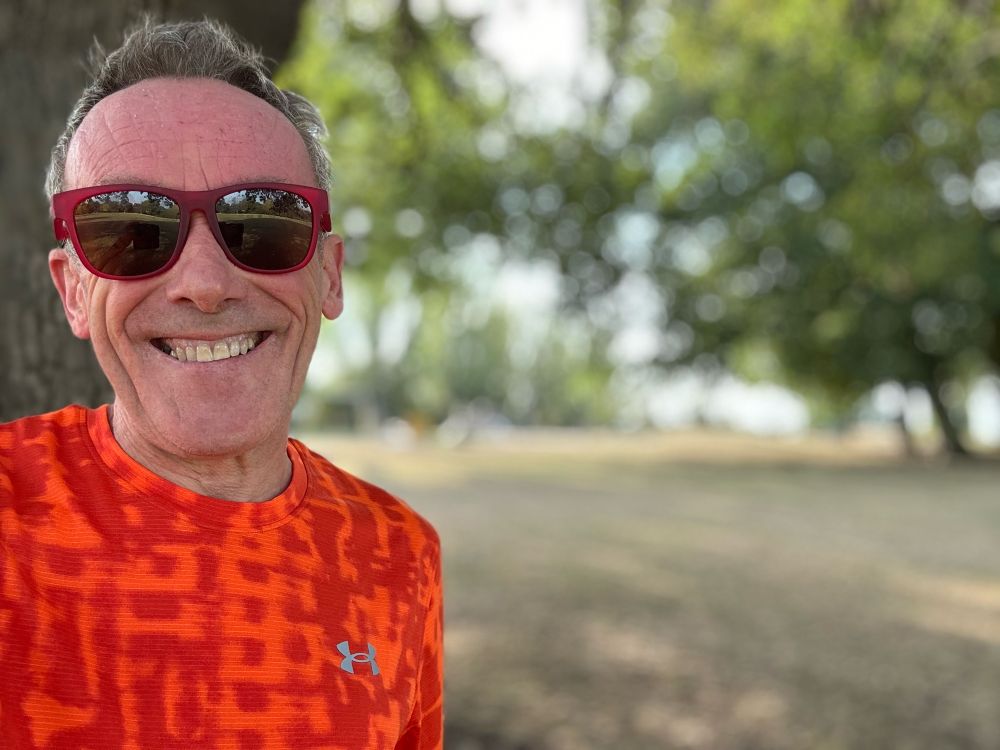 Smiling runner, hair a mess, grinning into the camera with red sunglasses and orange and terracotta t shirt.