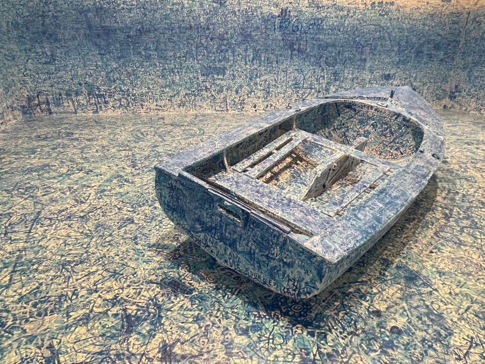 A largely blue boat which is a participatory piece of art by Yoko Ono called ‘Add Colour (Refugee Boat)’ that speaks to the current refugee crisis and whilst at the Tate in 2024 allowed people to add their own comments both onto the boats and in the room it was exhibited.