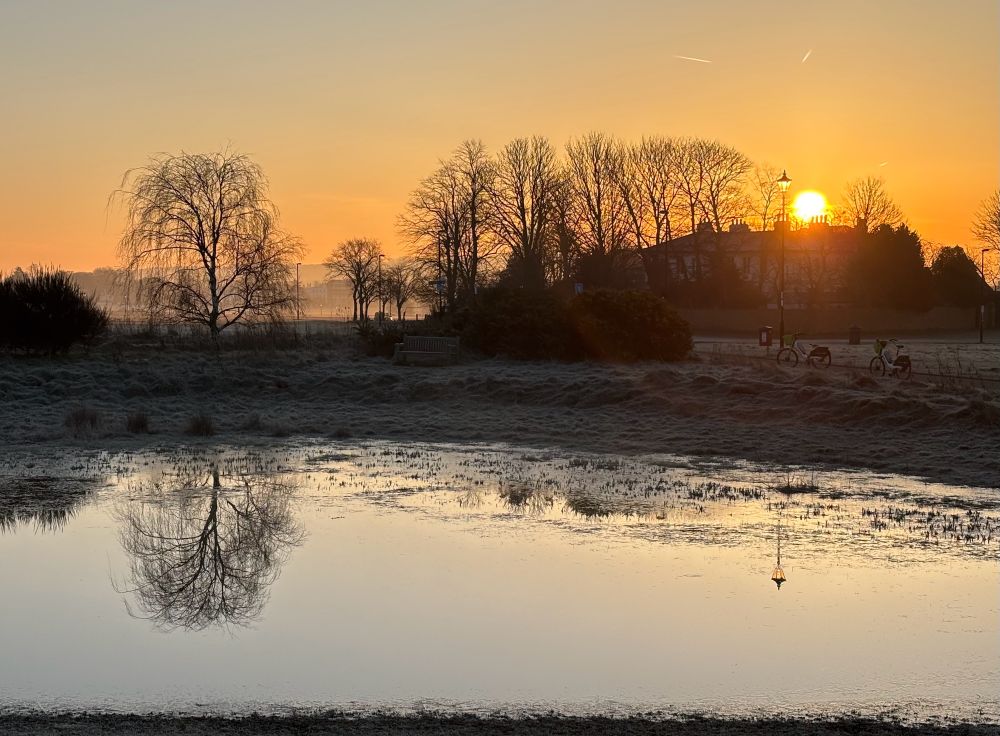A small group of buildings and trees surrounded by frosty grass with the sun rising above.  Almost all is reflected in the pond.