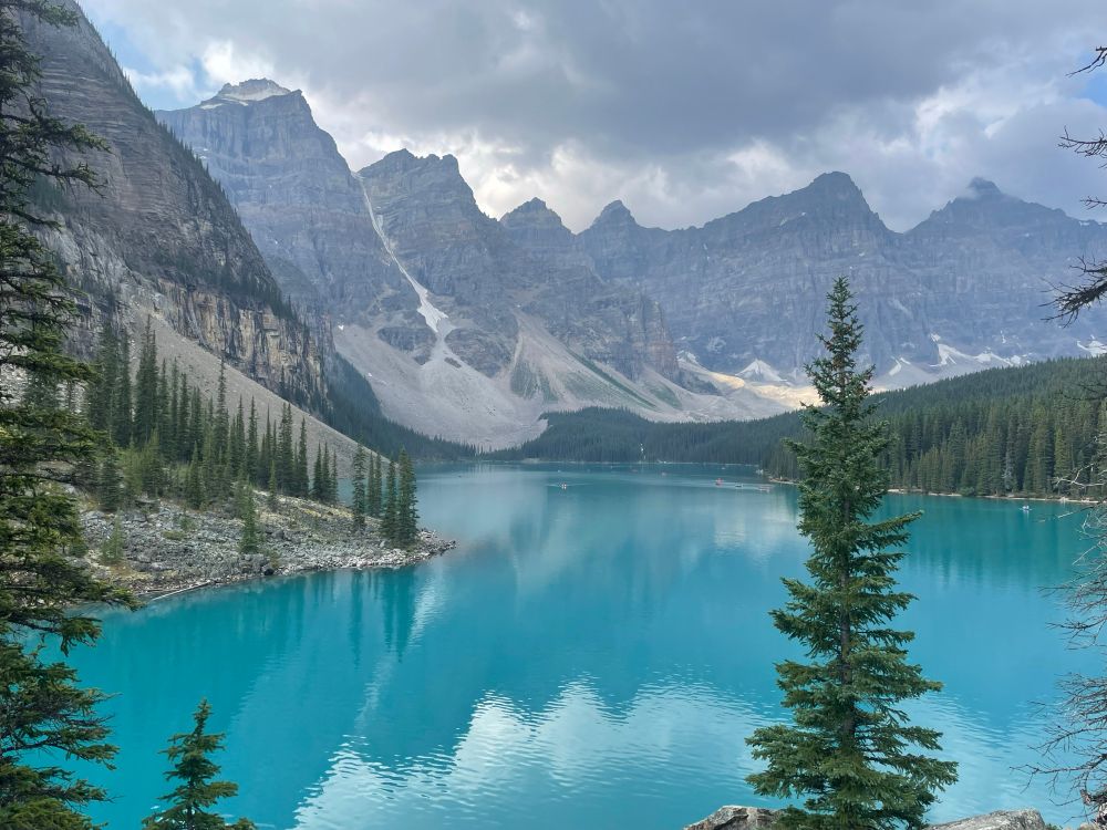 A bright blue lake surrounded by peaked mountains and glaciers. The lower slopes of the areas not covered by ice there are conifers.  