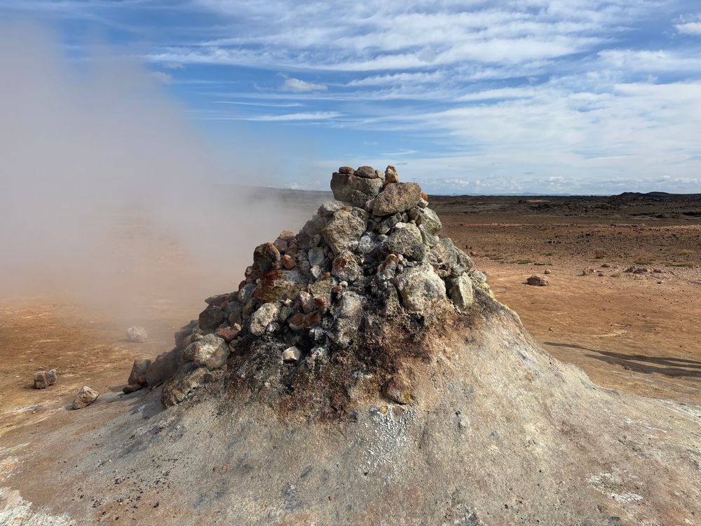 Steam pouring out of the left of a conical mound. Behind is sandy coloured volcanic debris and blue skies.  It is fortunate that there is no smell with this as the stench of sulphur would be overpowering. 