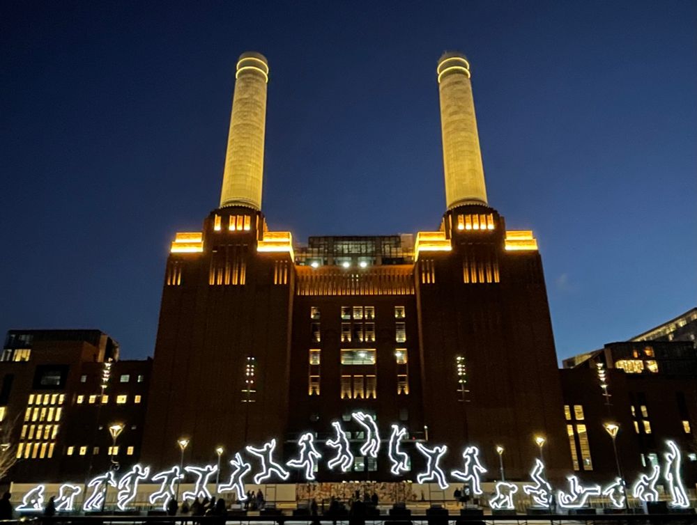 A large former power station now flats and a shopping centre. Its former chimneys are lit as are upper bits of the building. In front, about 100 metres away is a light installation called Run Beyond by Angelo Bonello, it was there for the Winter Lights Festival in 2022.