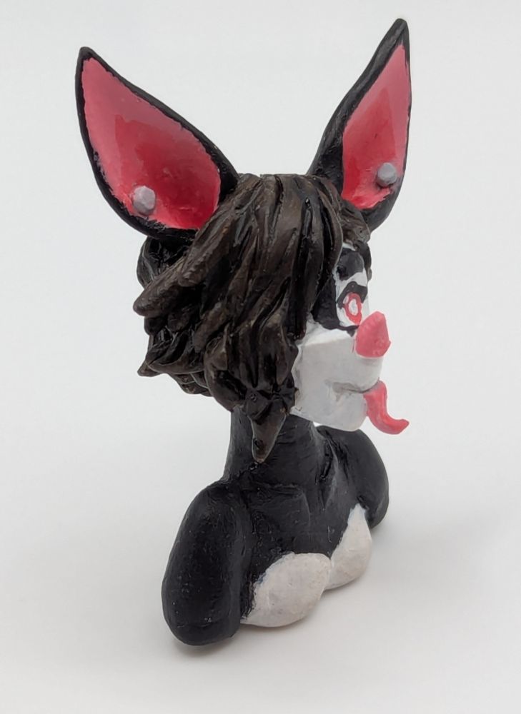 A polymer clay of an anthopormophic bat. She has her tongue sticking out in a playful expression, her bangs cover one eyes. She is a majority black and white, with pink inner ears, nose and eyes.