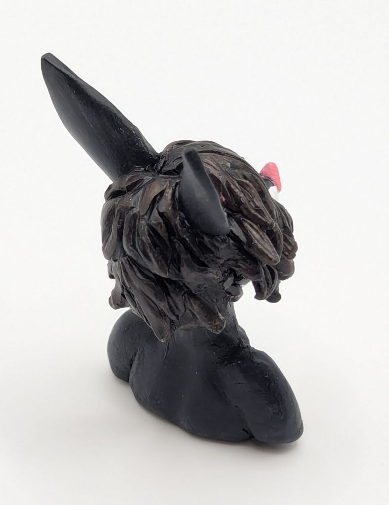 A polymer clay of an anthopormophic bat. She has her tongue sticking out in a playful expression, her bangs cover one eyes. She is a majority black and white, with pink inner ears, nose and eyes.