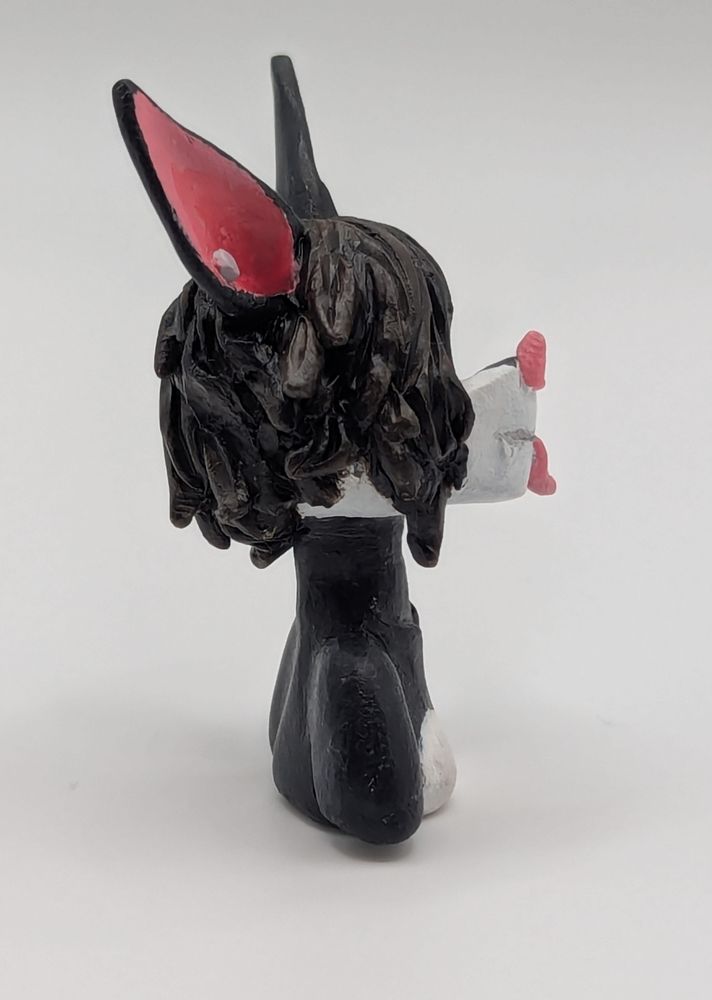 A polymer clay of an anthopormophic bat. She has her tongue sticking out in a playful expression, her bangs cover one eyes. She is a majority black and white, with pink inner ears, nose and eyes.