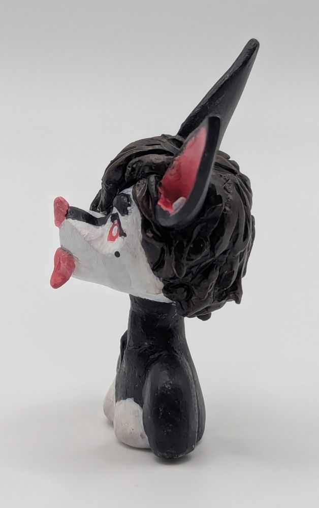 A polymer clay of an anthopormophic bat. She has her tongue sticking out in a playful expression, her bangs cover one eyes. She is a majority black and white, with pink inner ears, nose and eyes.