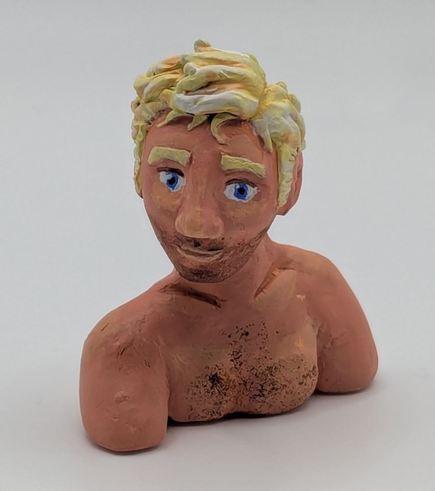A polymer clay bust of a Caucasian blonde man. He has blue eyes and slightly unkept but combed hair. He is leaning slightly forward. He has stubble and chest hair.