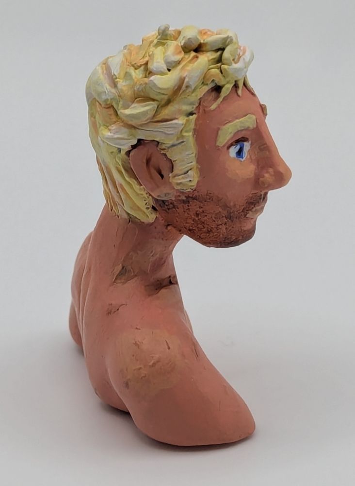 A polymer clay bust of a Caucasian blonde man. He has blue eyes and slightly unkept but combed hair. He is leaning slightly forward. He has stubble and chest hair.
