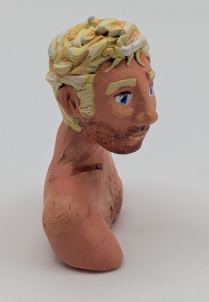A polymer clay bust of a Caucasian blonde man. He has blue eyes and slightly unkept but combed hair. He is leaning slightly forward. He has stubble and chest hair.