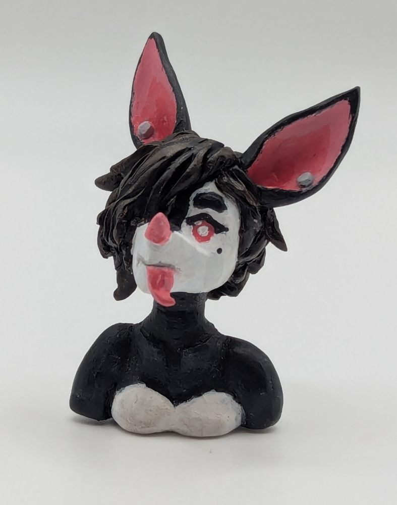 A polymer clay of an anthopormophic bat. She has her tongue sticking out in a playful expression, her bangs cover one eyes. She is a majority black and white, with pink inner ears, nose and eyes.