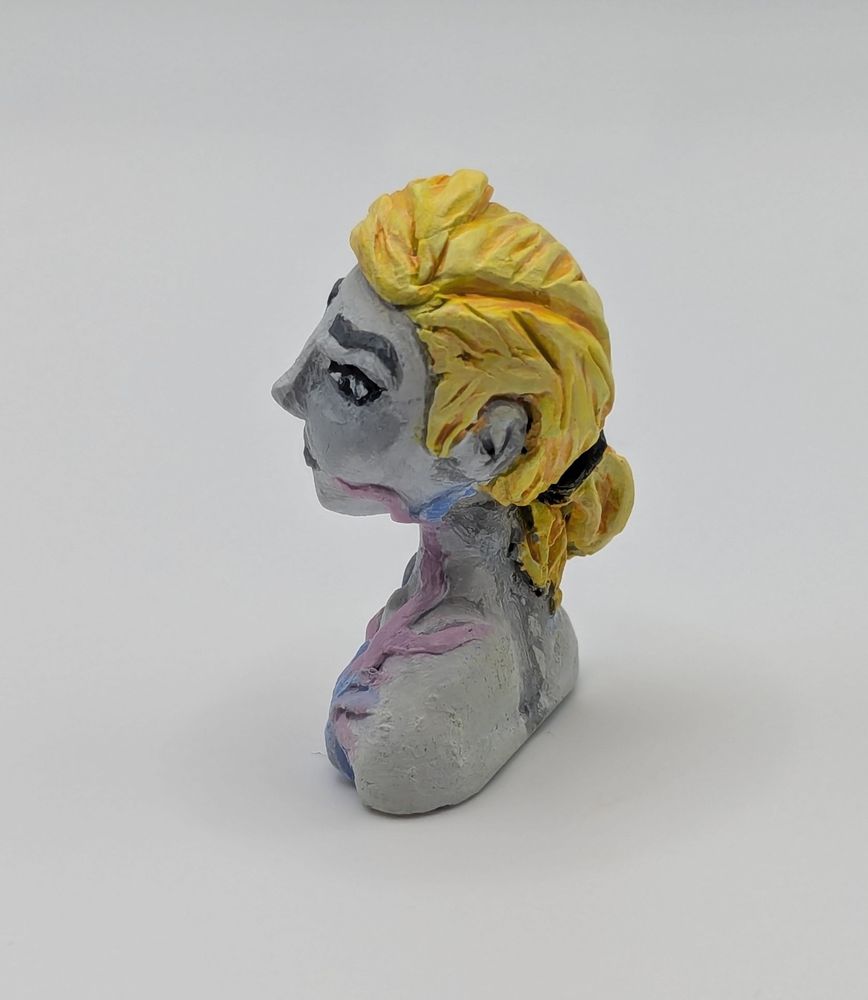 A clay bust of a human female. She has a limited greyscale pallet save for her blonde hair and a strange pink and blue biological growth on her chest.