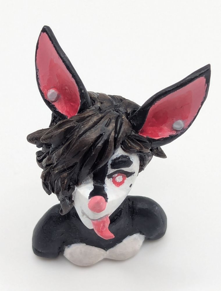 A polymer clay of an anthopormophic bat. She has her tongue sticking out in a playful expression, her bangs cover one eyes. She is a majority black and white, with pink inner ears, nose and eyes.