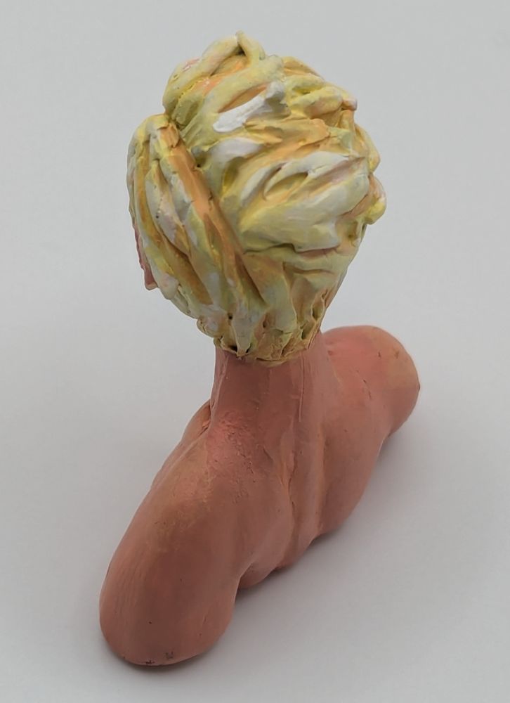 A polymer clay bust of a Caucasian blonde man. He has blue eyes and slightly unkept but combed hair. He is leaning slightly forward. He has stubble and chest hair.