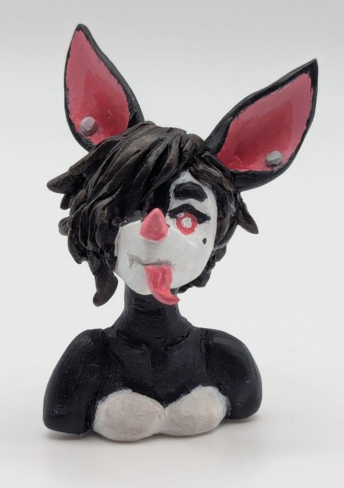 A polymer clay of an anthopormophic bat. She has her tongue sticking out in a playful expression, her bangs cover one eyes. She is a majority black and white, with pink inner ears, nose and eyes.