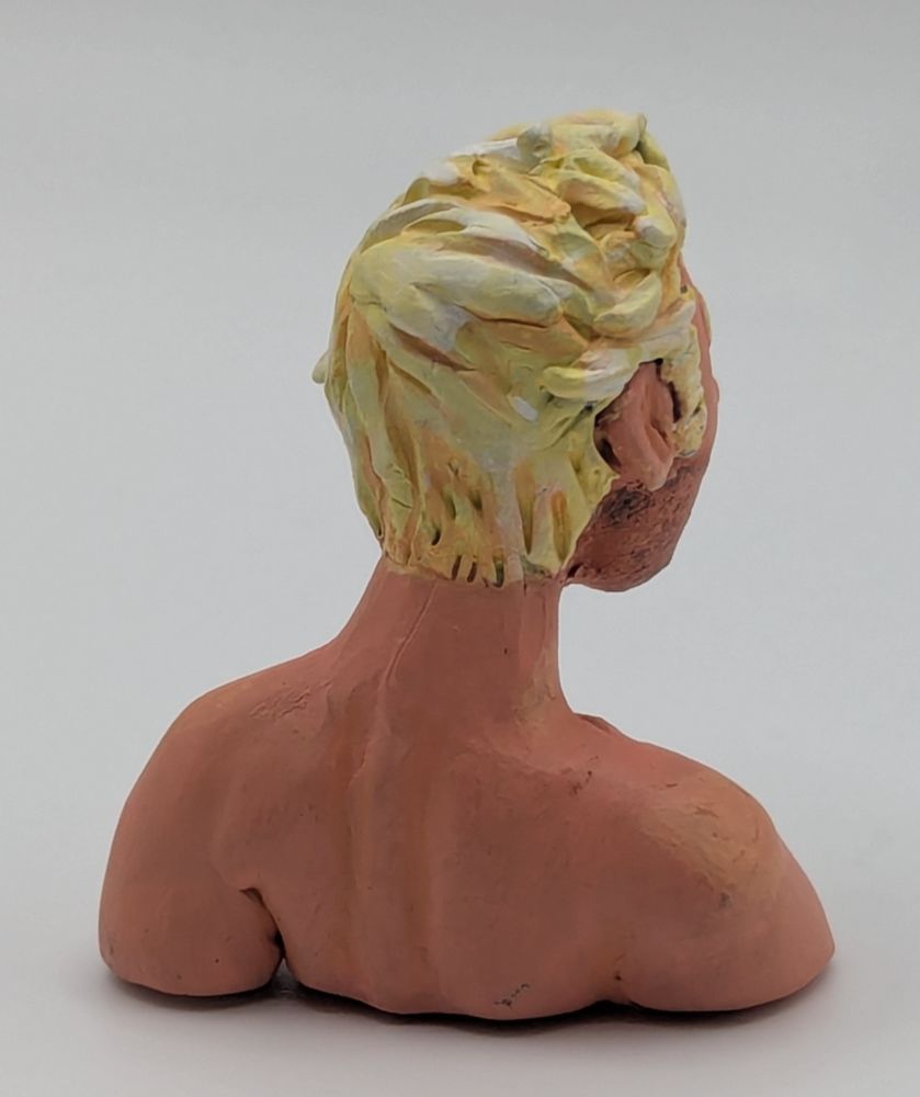 A polymer clay bust of a Caucasian blonde man. He has blue eyes and slightly unkept but combed hair. He is leaning slightly forward. He has stubble and chest hair.