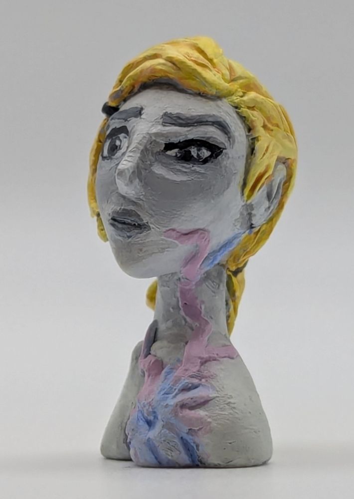 A clay bust of a human female. She has a limited greyscale pallet save for her blonde hair and a strange pink and blue biological growth on her chest.