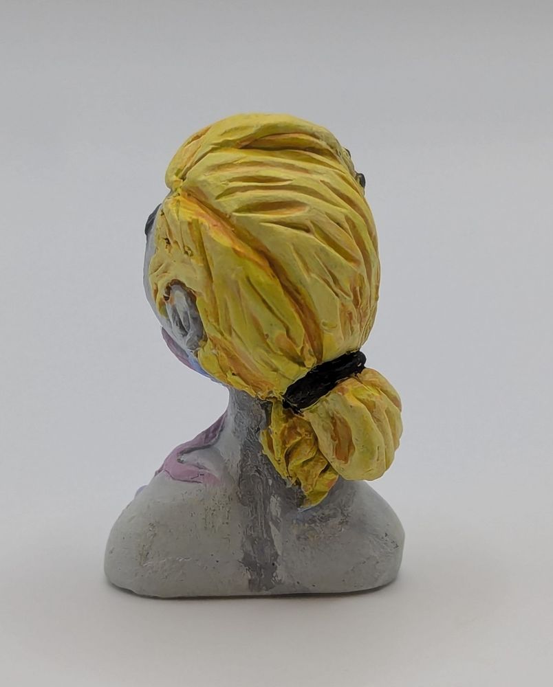 A clay bust of a human female. She has a limited greyscale pallet save for her blonde hair and a strange pink and blue biological growth on her chest.