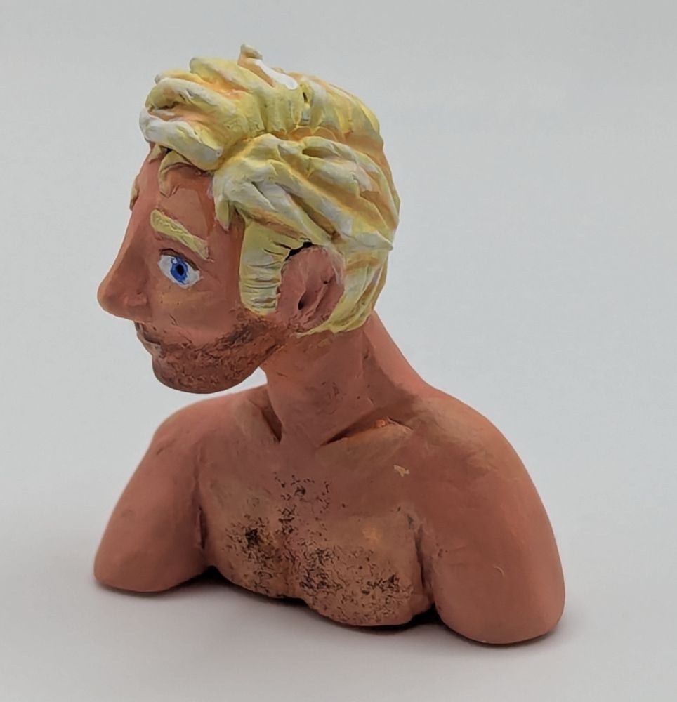 A polymer clay bust of a Caucasian blonde man. He has blue eyes and slightly unkept but combed hair. He is leaning slightly forward. He has stubble and chest hair.