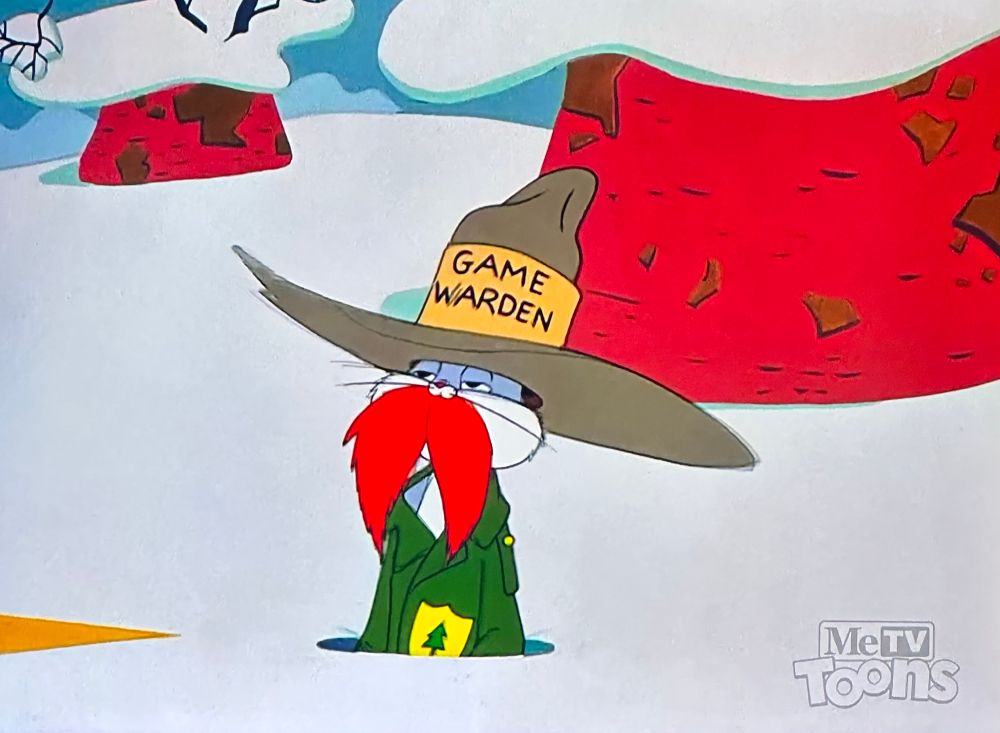 bugs bunny popping out of his hole in a game warden costume, he’s got a wide brimmed hat on that says “game warden” and a huge fake mustache and a green jacket on. 