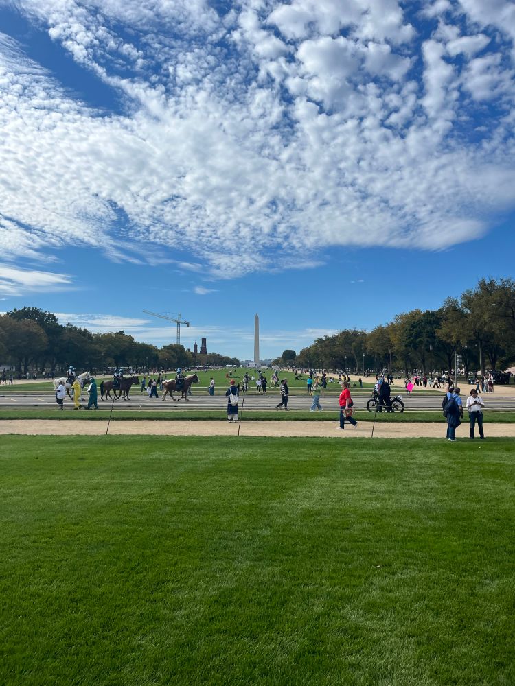 A blue sky with white clouds above the Washington Monument and the green grass and tan gravel paths of the National Mall with people milling about in the foreground
