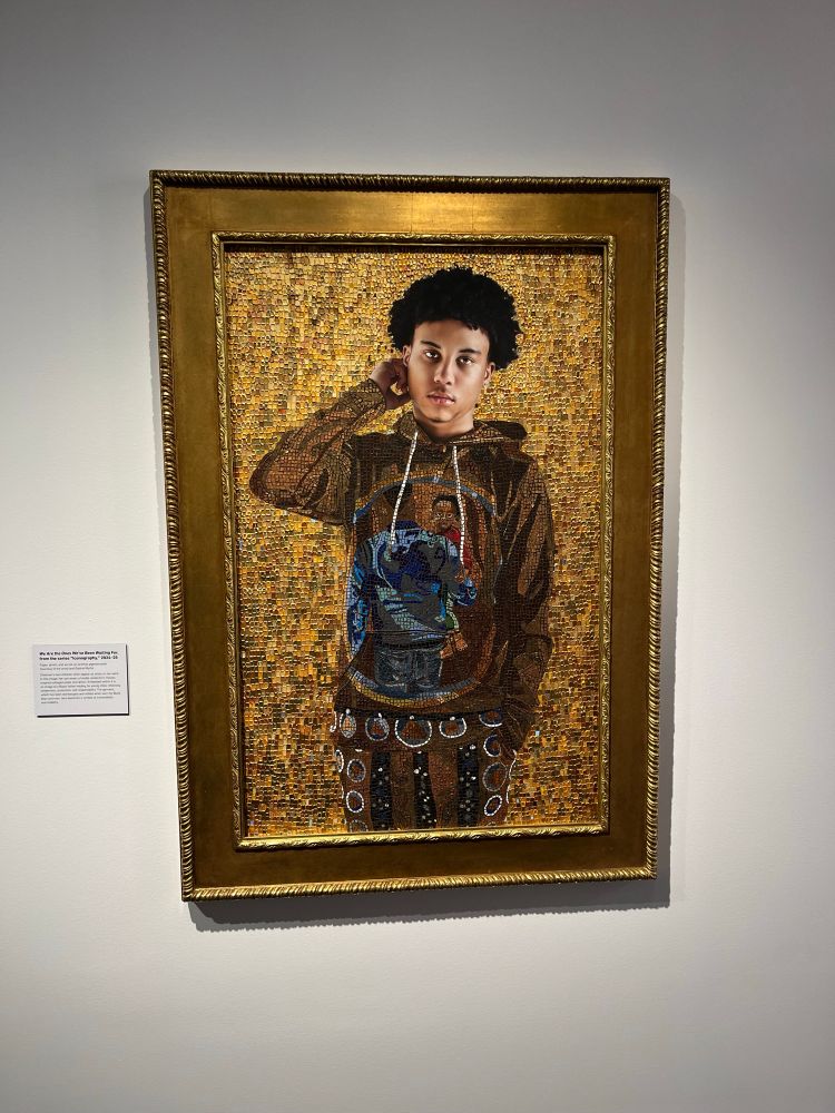A portrait in a gold frame on a white wall featuring a Black teenage boy in a hoodie sweatshirt that has an image of a pair of male figures on it. The background of the portrait is a gold mosaic