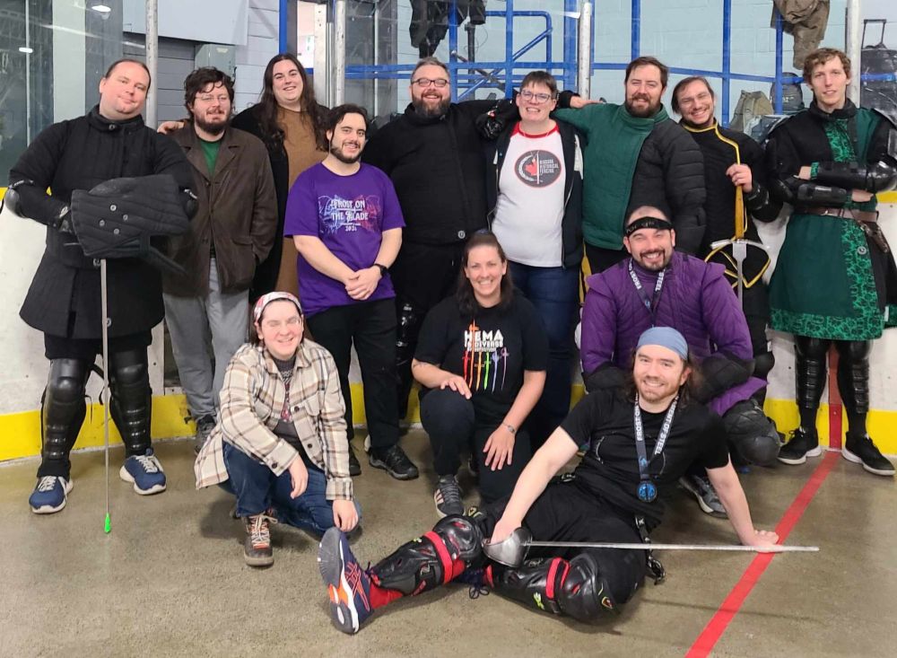 A group of fencers standing, kneeling, and sprawling. Some of us are wearing padded jackets or holding sparring swords, but some are in regular clothes too. All of us are pretty gassed from fencing or cheering each other on. Everyone's smiling because it was hella fun, and each victory a group effort.