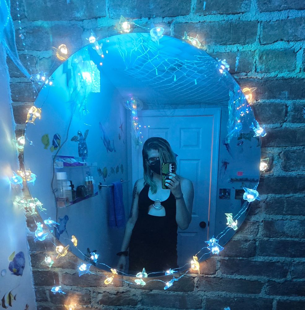 Goth girl in a black dress in a room lit dimly with blue light and fairie lights. she is framed by a circular mirror 