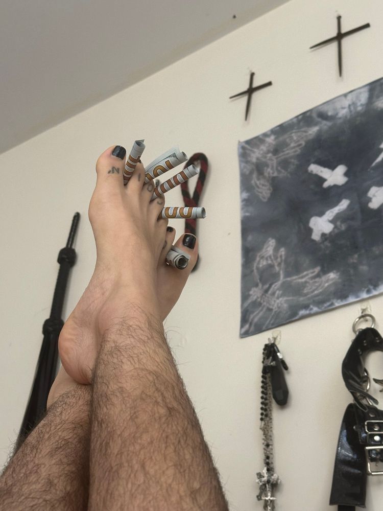 Five hundred dollar bills are rolled and stuffed in between a white persons toes. Their legs can be seen and are hairy. In background hangs many impact toys, two crucifixes and a blue banner with tied hands and more crucifixes.