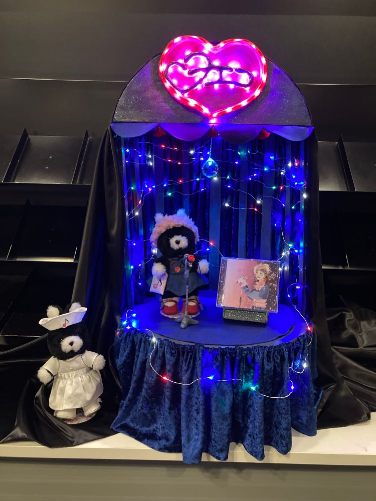 ネオン装飾のされた青いぬいぐるみ用の小さなステージの上に黒いうさぎのマスコットのおちょこちゃんがおしゃれをして立ってる。ステージの横にはナース服を着たもう1匹のおちょこちゃんが佇んでる。