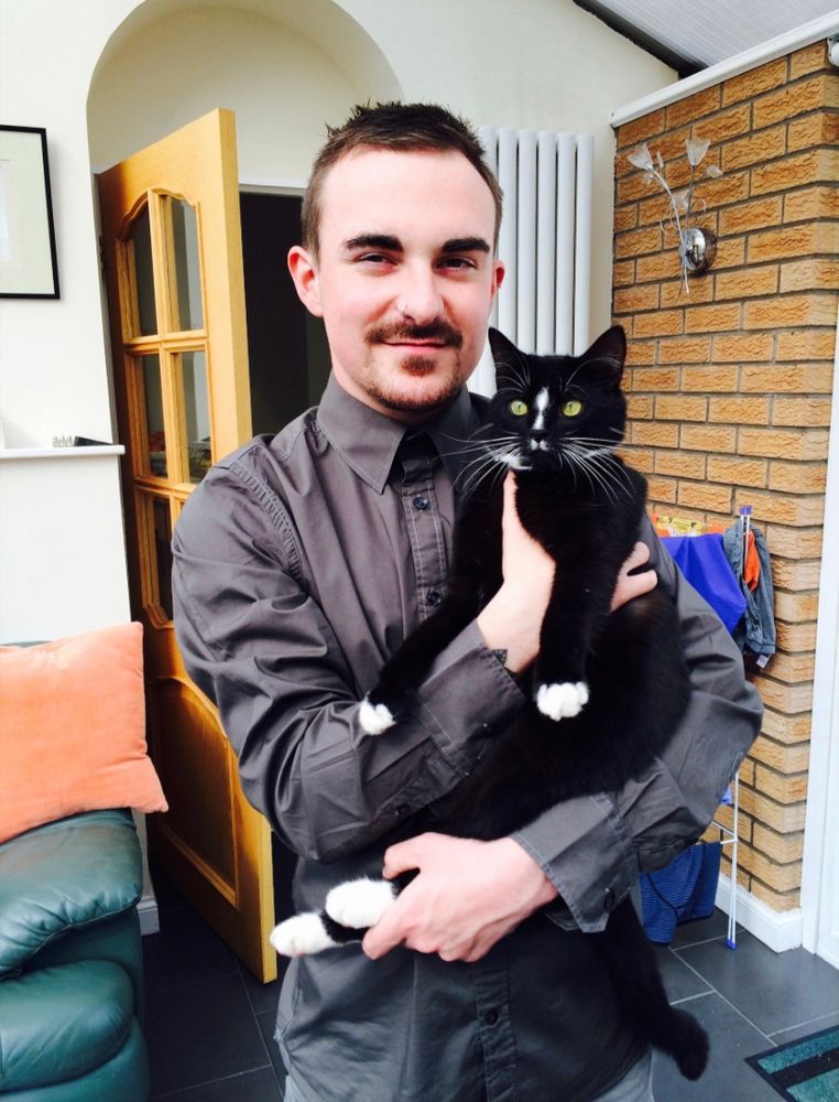 A man in a grey shirt holding a black cat with white paws, smiling in a cosy indoor setting.