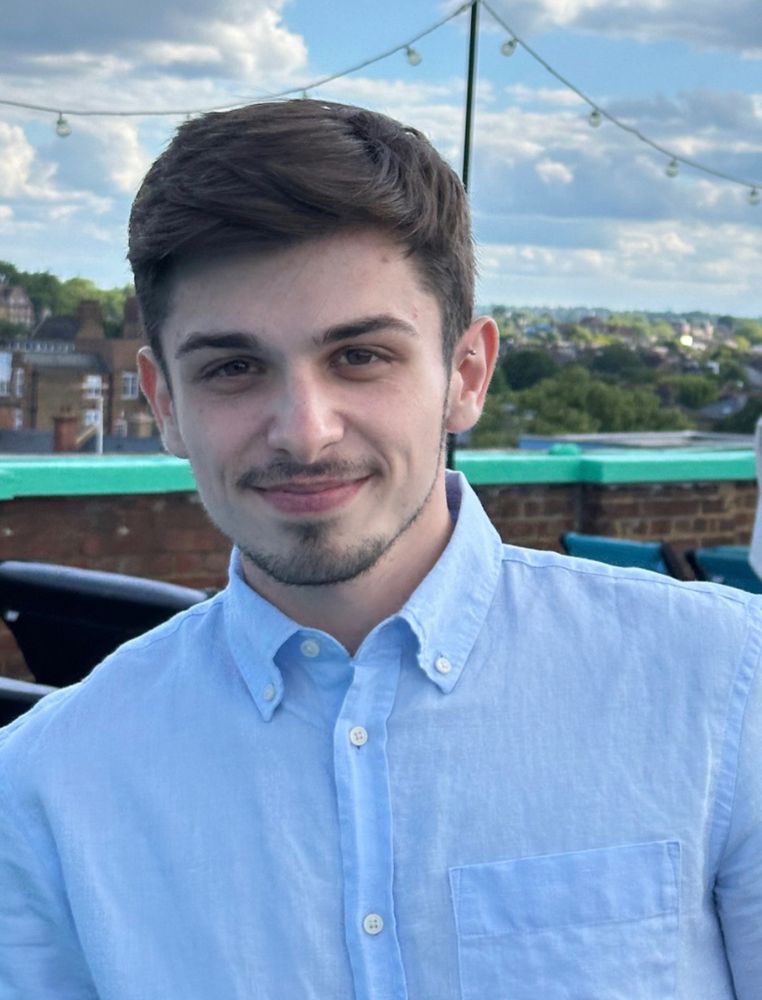  A man in a light blue shirt smiles at the camera on a rooftop with a cityscape in the background.