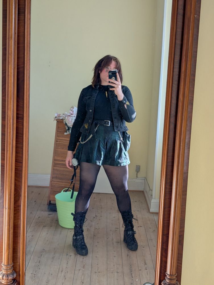 an alt woman wearing a black denim jacket, a skirt, and boots 
