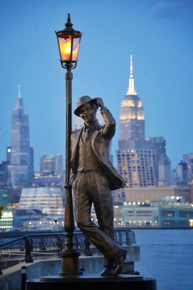 In blue twilight, a statue of Frank Sinatra leaning against a lamppost, tipping his hat. The Empire State Building is lit up in the background.