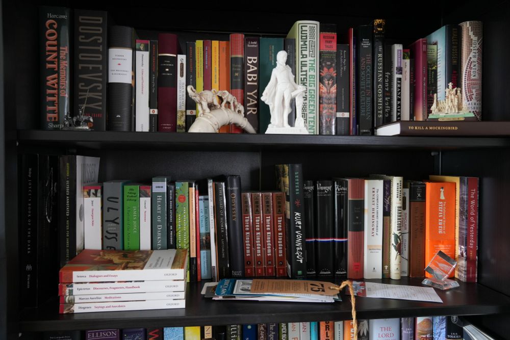 Top shelf is a selection of books. Mostly Russian literature with some assorted other literature at the right side. There are sculptures of Don Quixote, elephants on a horn, Mozart, and Sagrada Família in front of the books on the top shelf.
The second shelf is literature from all over the world. There are bookmarks in the middle. A glass seethrough cube with a base that says "U.S. Cap tol" [sic] stands at the front right.