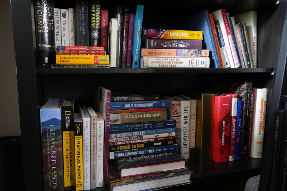 The top shelf is full of scientific/nerdy books with some folk tales in the middle. There is a small selection of phrasebooks and language translation books in the front left, specifically for Latin American Spanish, Russian, and French. At the bottom of these language books is an old box manual for Warcraft 3.
The second shelf is full of books on nature, outdoors, physical training (especially for hiking/mountaineering), more political pop books, and an assortment of oddities like a book about professional criminals in america dating back to 1886 by Byrnes.