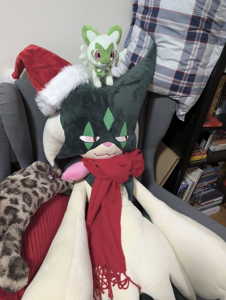 A Meowscarada plush sitting in an armchair. There is a red scarf tied around the Meowscarada and a santa hat on the left mask bit poking out of Meowscarada.  A leopard plush on a red pillow lies to the left of it. A Sprigatito sits on the Meowscarada's head. A library full of books is seen to the right.