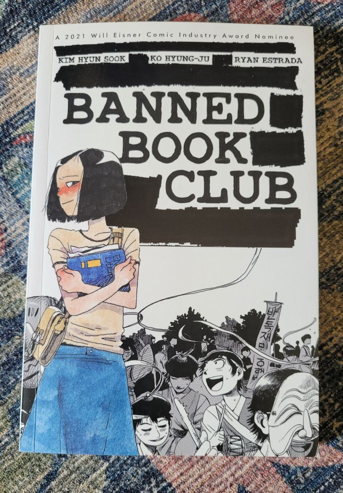 Photo of graphic novel Banned Book Club by Kim Hyun Sook, Ko Hyung-Ju, and Ryan Estrada on a muted blue and pink floral rug. Cover of the book is a drawing of Hyun Sook clutching what looks like a binder with papers sticking out and with a group of protestors behind her. 
