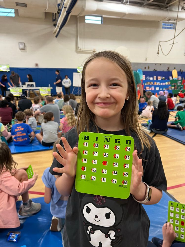 Student smiles holding BINGO card. Students and families sit on mats in the gym eating pizza before and while playing BINGO
