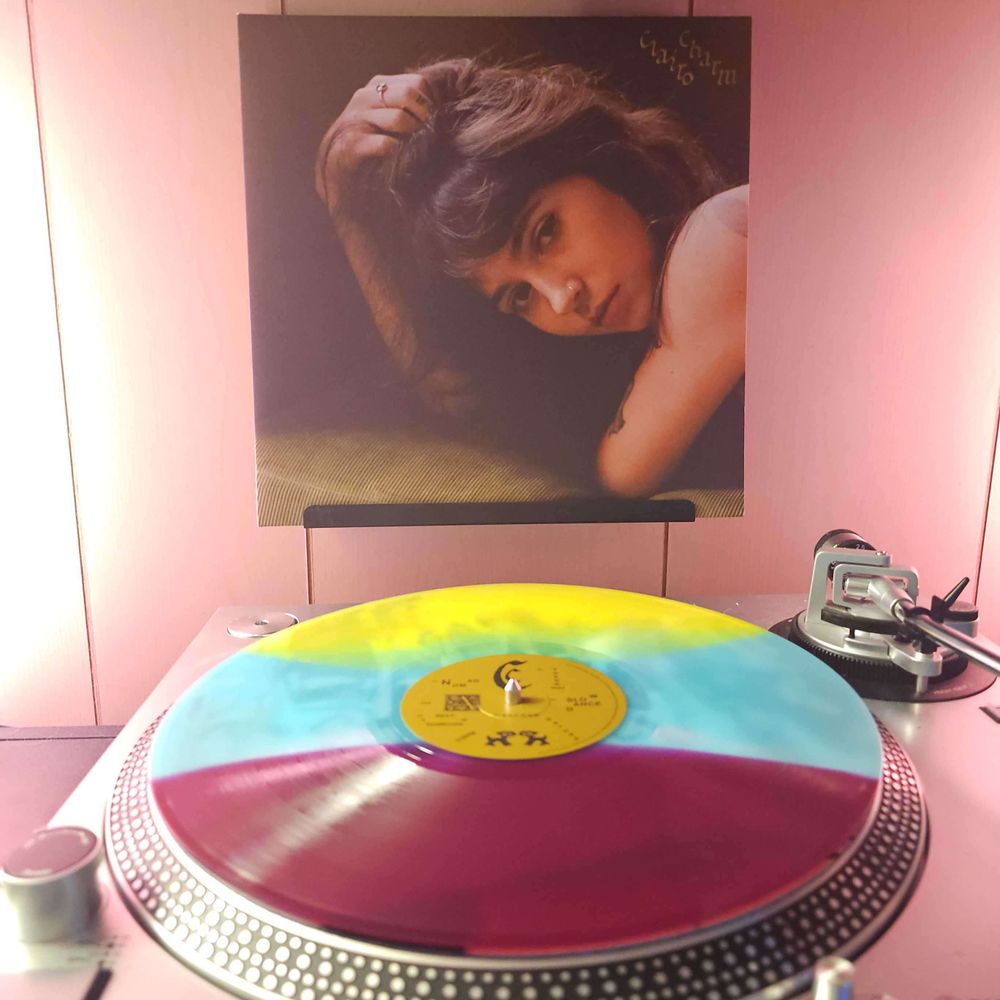 A Yellow, Light Blue & Purple Striped vinyl record sits on a turntable. Behind the turntable, a vinyl album outer sleeve is displayed. The front cover shows Clairo from the shoulders up. She is laying on a cushion, with her right hand grabbing hair on top of her head as she looks at the camera.