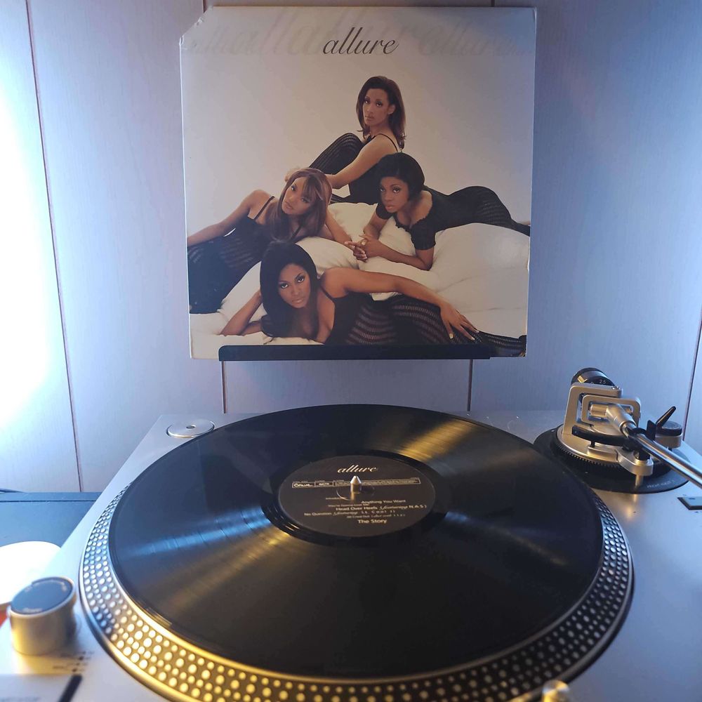 A vinyl record sits on a turntable. Behind the turntable, a vinyl album outer sleeve is displayed. The front cover shows the 4 members of allure laying around on a large amount of white bedding. Each member is wearing a long black negligee dress that is see through in some areas.